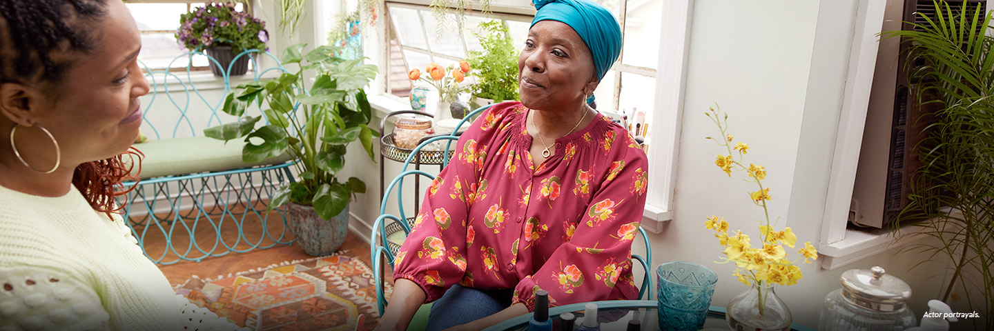A Black woman in her mid-50s wearing a teal headscarf is sitting at a glass table in a brightly lit sunroom. She is with her friend, a Black woman in her late 40s with long braids and hoop earrings who is giving her a manicure. Colorful nail polish bottles are on the table. Not actual patients.